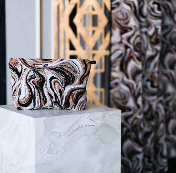 Косметичка Feiler Hermitage MURANO NERO с чёрно-бело-медными мраморными разводами и петлёй-ручкой сбоку стоит на белом каменном постаменте, справа штора того же рисунка