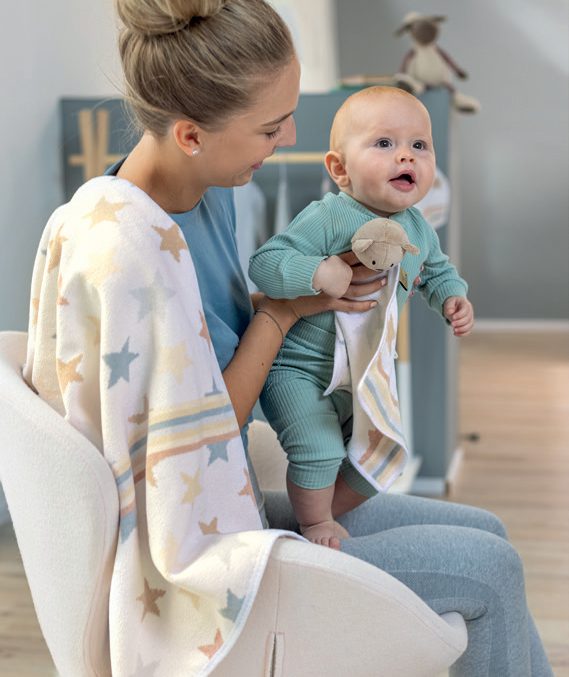 Детское одеяло Feiler Mini STARS & STRIPES 75x100 с бежевыми и серыми звёздами и полосатой каймой перекинуто через плечо матери; она держит малыша, у малыша в руках салфетка с игрушкой той же коллекции